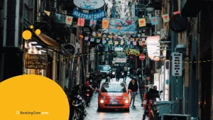 Calle estrecha en Roma con banderines, letreros y un Fiat rojo entre peatones.
