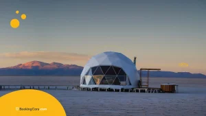 Domo geodésico de glamping junto a lago con montañas al atardecer.