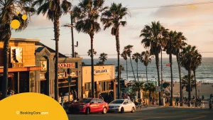 Calle costera con palmeras, autos, comercios frente al mar y atardecer.